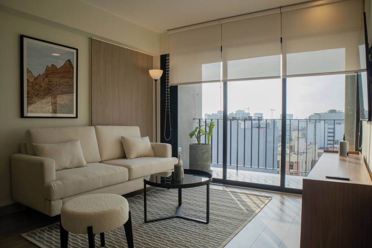 Uma sala de estar moderna com um sofá, uma mesa de centro e vista para o horizonte urbano através de grandes janelas.
