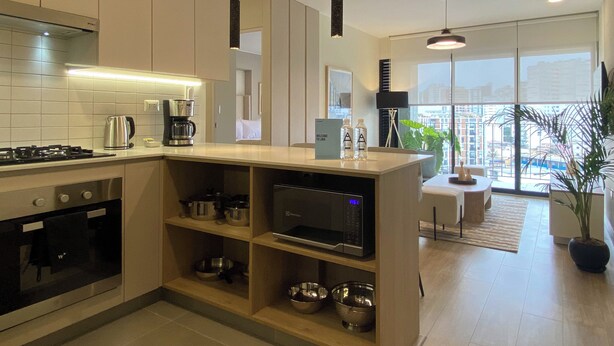A modern kitchen with a gas stove, microwave, and coffee maker. There is a dining area with a table and chairs, and a large window offering a city view. - Pod by Wynwood House (Lima)
