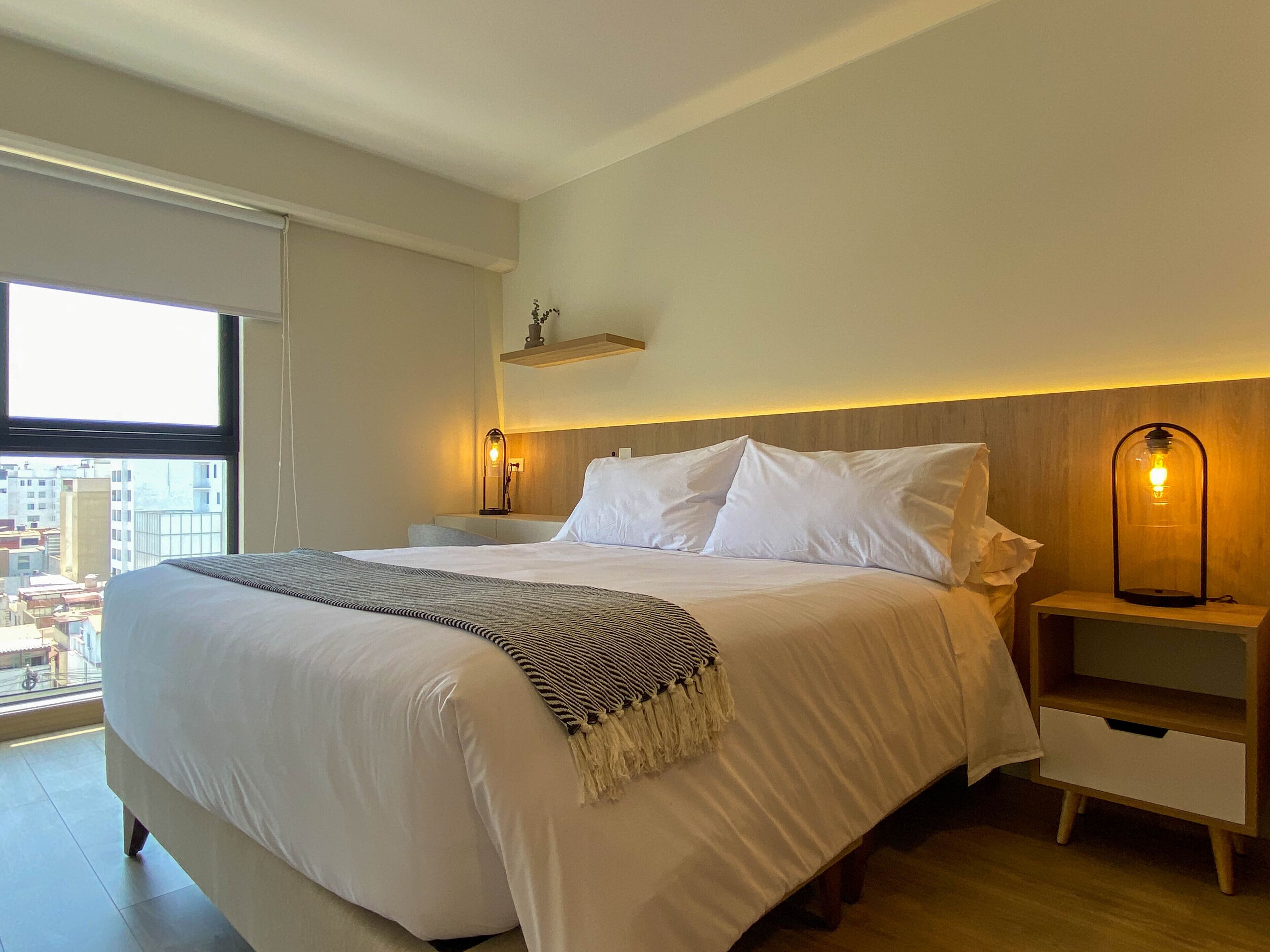 A modern hotel room with a large bed, a bedside table, a nightstand, and a view of the city through a large window.