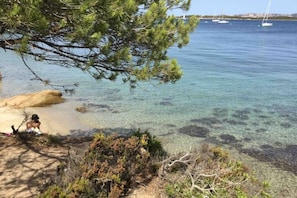 A beach with clear water, a rocky shore, and boats in the distance.