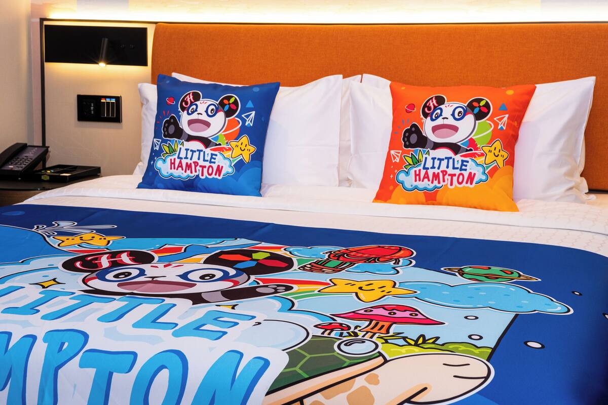 A hotel room with a bed featuring a colorful bedspread with 'Little Hampton' text and cartoon characters, two pillows with 'Little Hampton' text, a bedside table with a phone, and a wall-mounted control panel.