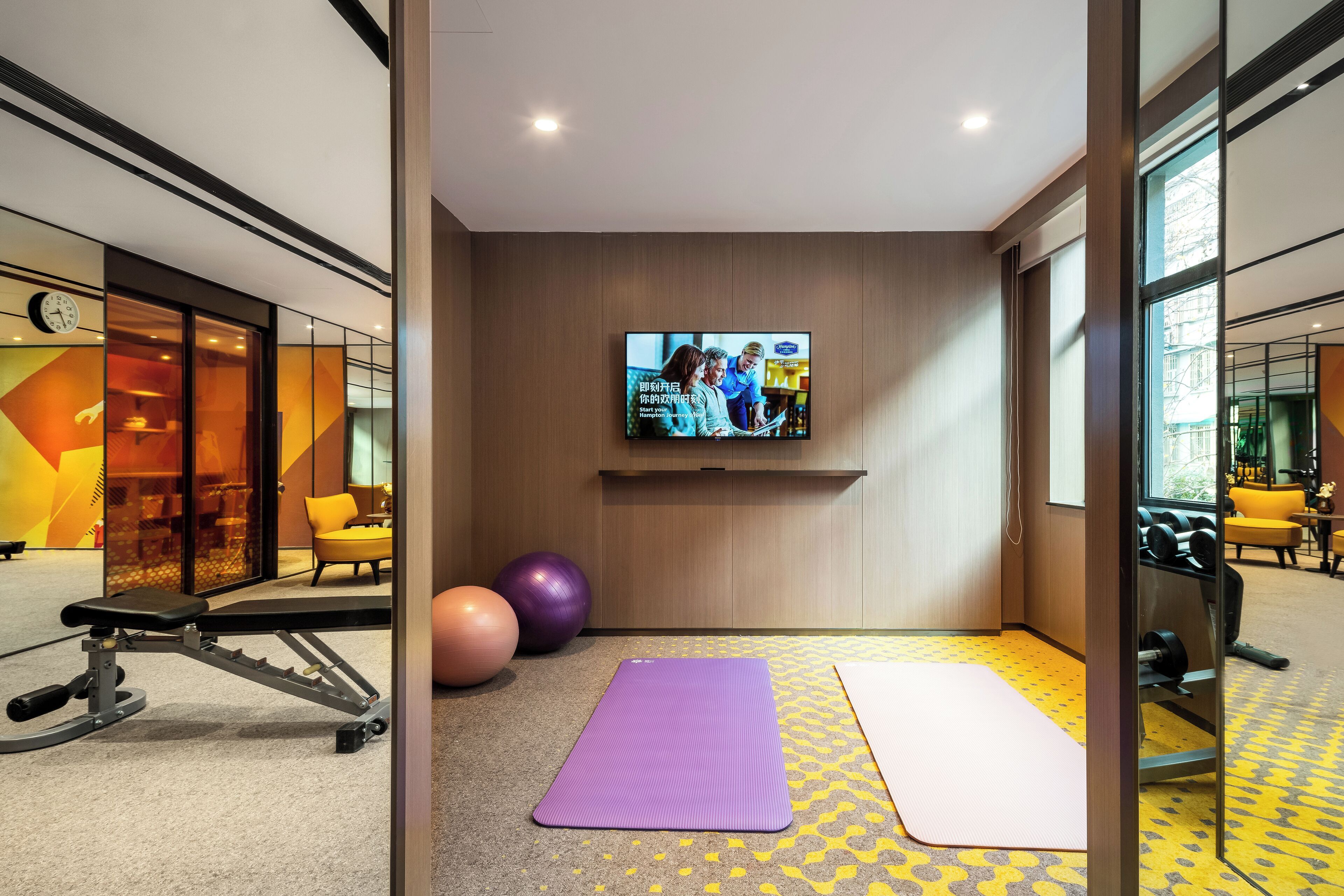 A modern gym with exercise equipment, a large TV screen, and a yellow patterned floor.