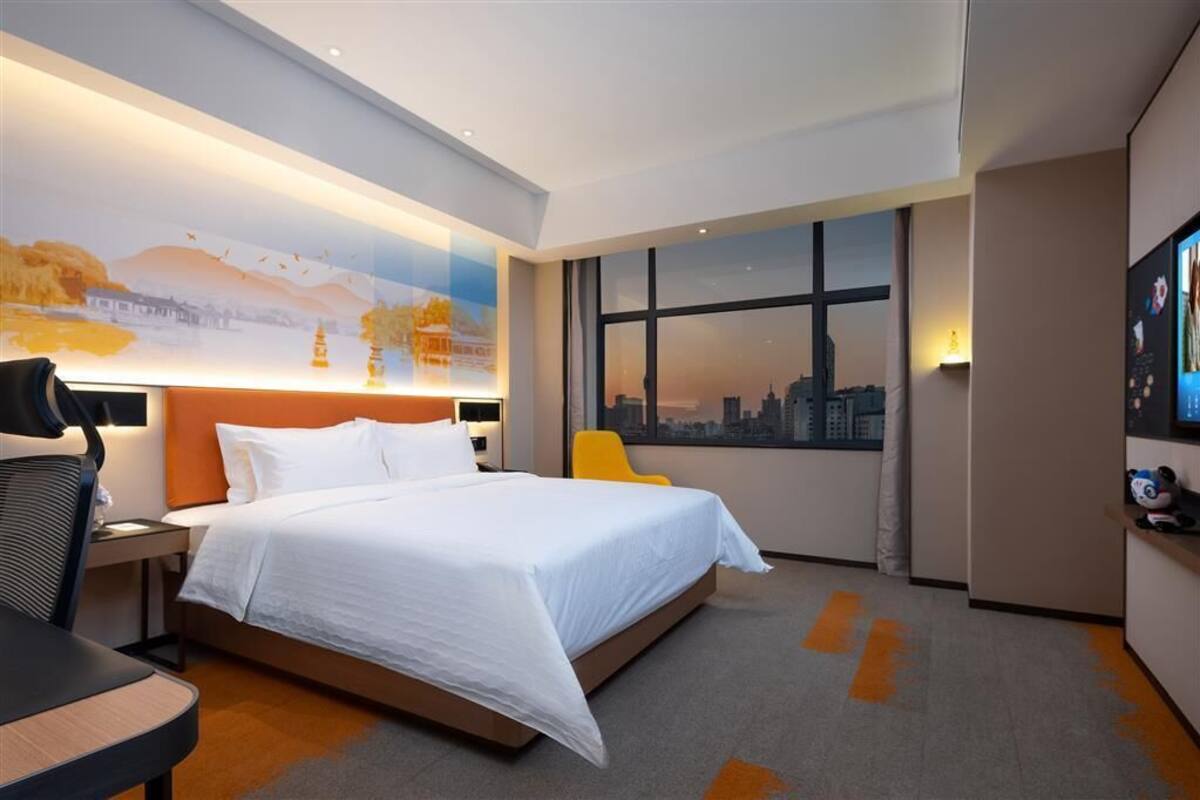 A modern hotel room with a large bed, a desk, a chair, and a cityscape view through the window.