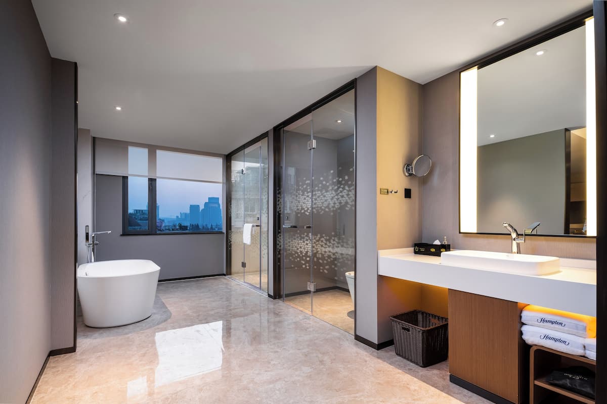A modern bathroom with a freestanding bathtub, a large mirror, and a city view through the window.