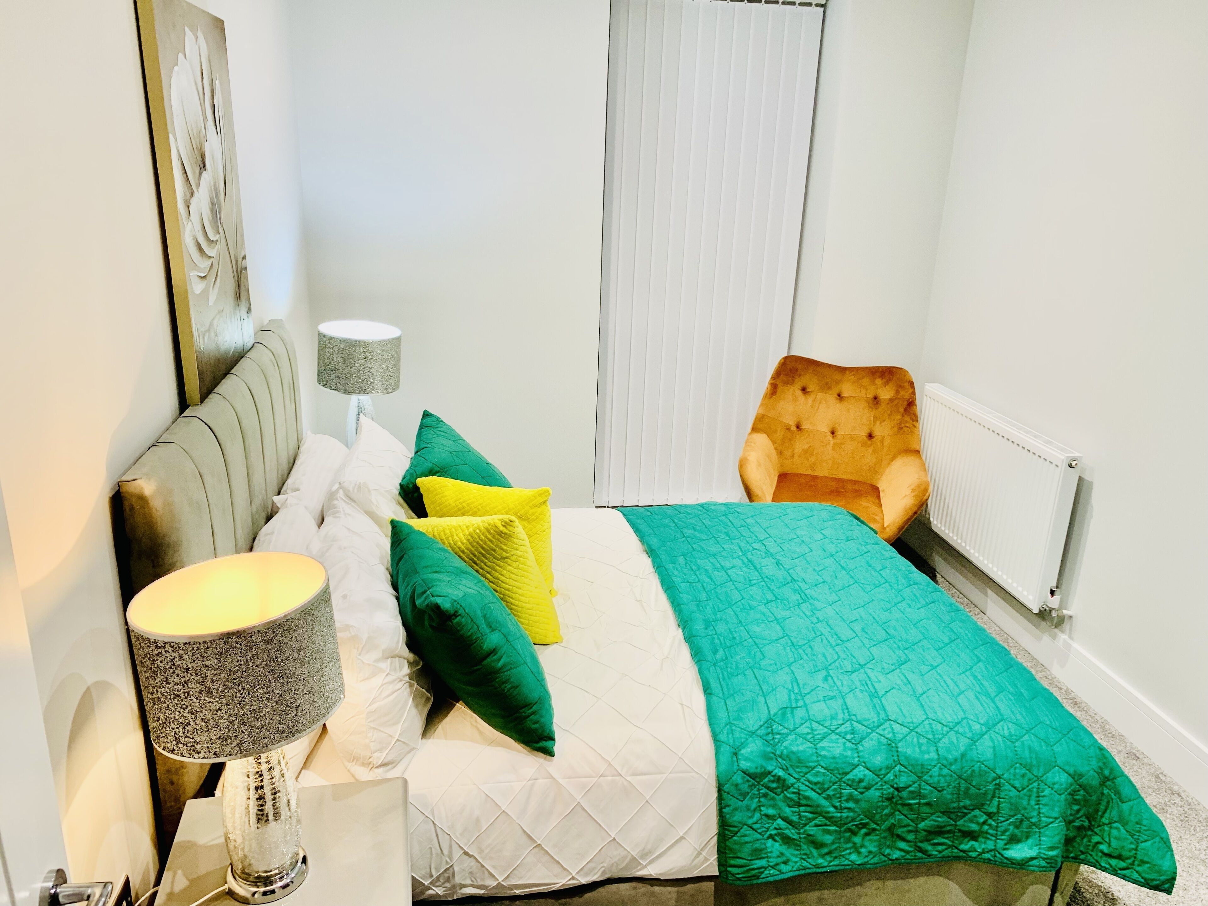 Ein ordentlich eingerichtetes Schlafzimmer mit einem Bett, Kissen, einem Sessel, einer Lampe und einer Heizung.