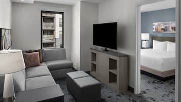 A modern hotel room with a grey sofa, a flat-screen TV, a bed, and a view of the city through a window.
