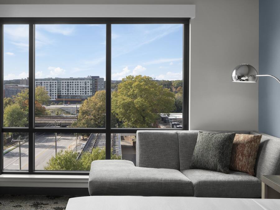 A modern hotel room with a large window offering a city view, a gray sofa, and a minimalist side table.