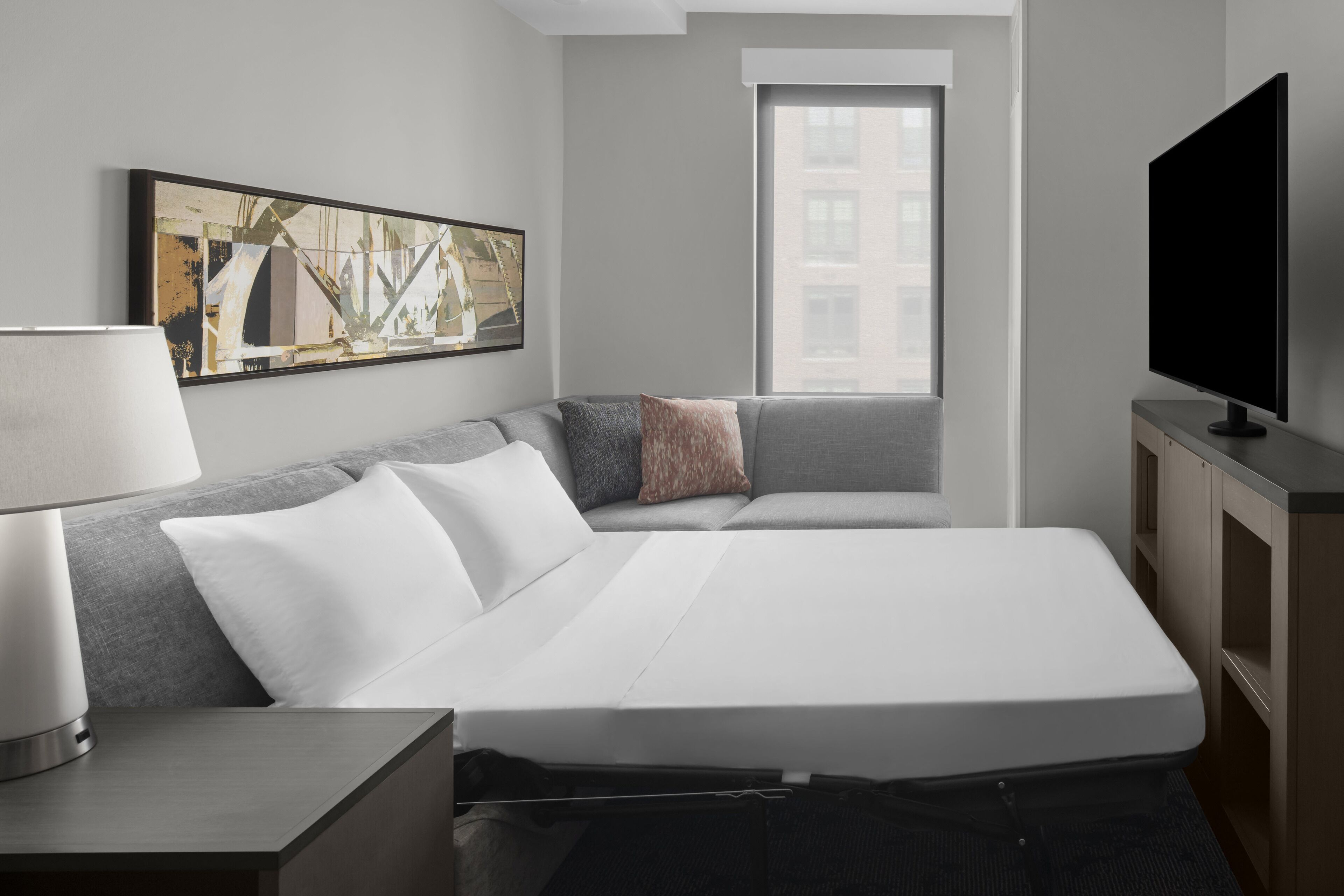 A modern hotel room with a bed, a TV, a desk, and a window with a view of buildings.