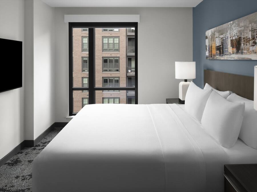 A hotel room with a large bed, a flat-screen TV mounted on the wall, a window with a city view, and a painting on the wall.