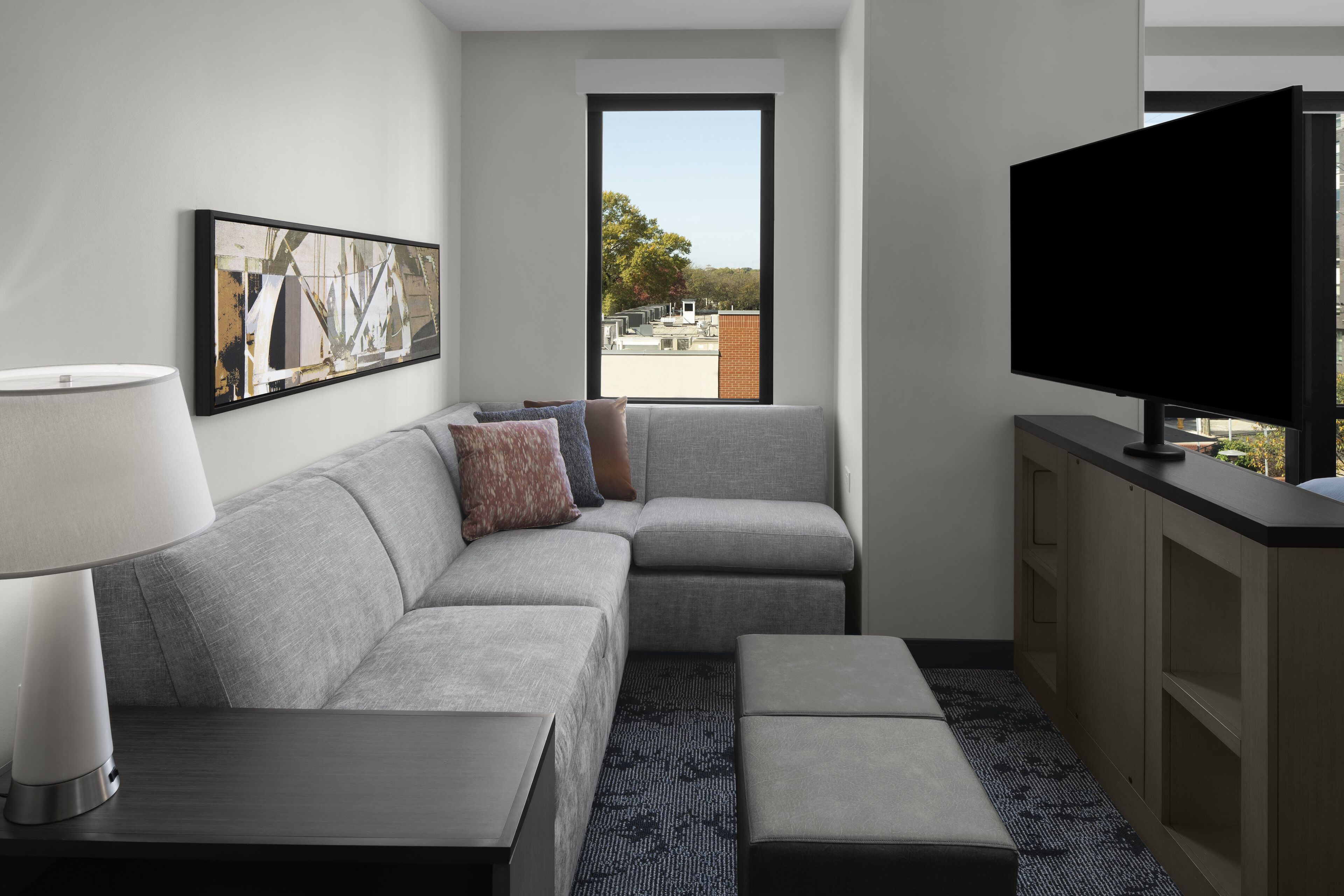 A modern hotel room with a grey sofa, a flat-screen TV, a side table, a lamp, and a large window with a view of buildings and trees.