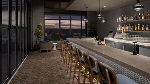 Een moderne bar met een lange toonbank, barstoelen en een uitzicht op de stad door grote ramen.