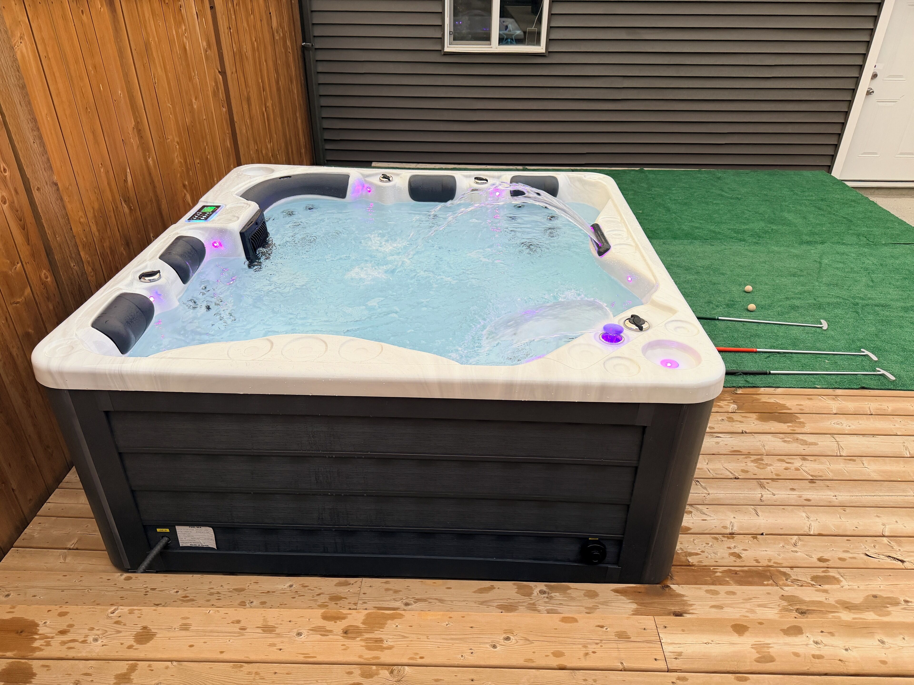 Un bain à remous doté d’un panneau de commande, entouré d’une terrasse en bois et d’une zone recouverte de gazon synthétique.