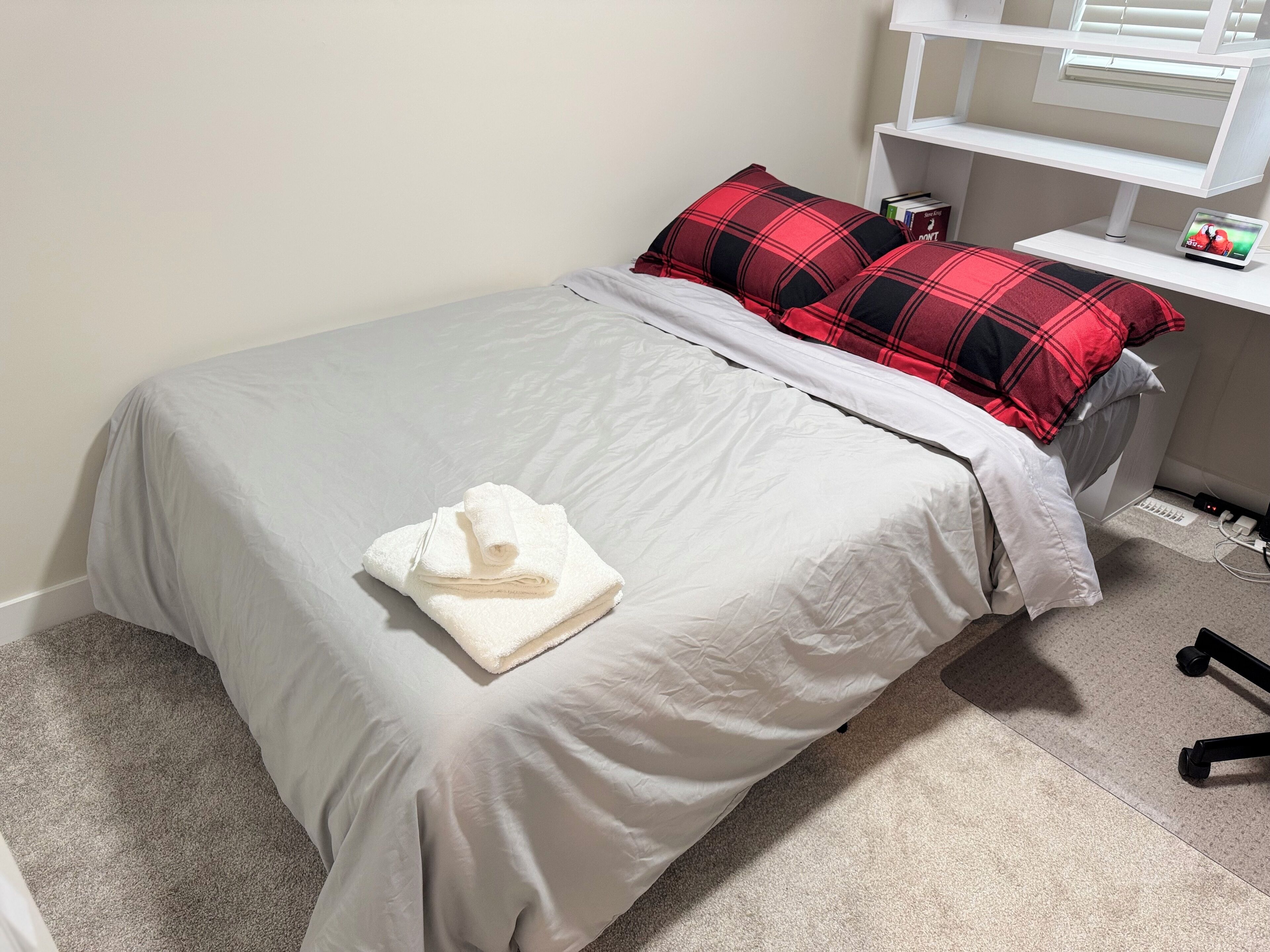 Un lit bien fait, avec une couette blanche et une serviette blanche pliée. On peut voir deux coussins à carreaux rouges et noirs sur le lit.