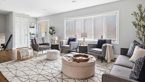A living room with a geometric patterned rug, a round ottoman, a grey sofa, and a grey armchair. - Spacious Home in Chicago Suburbs (Lombard)