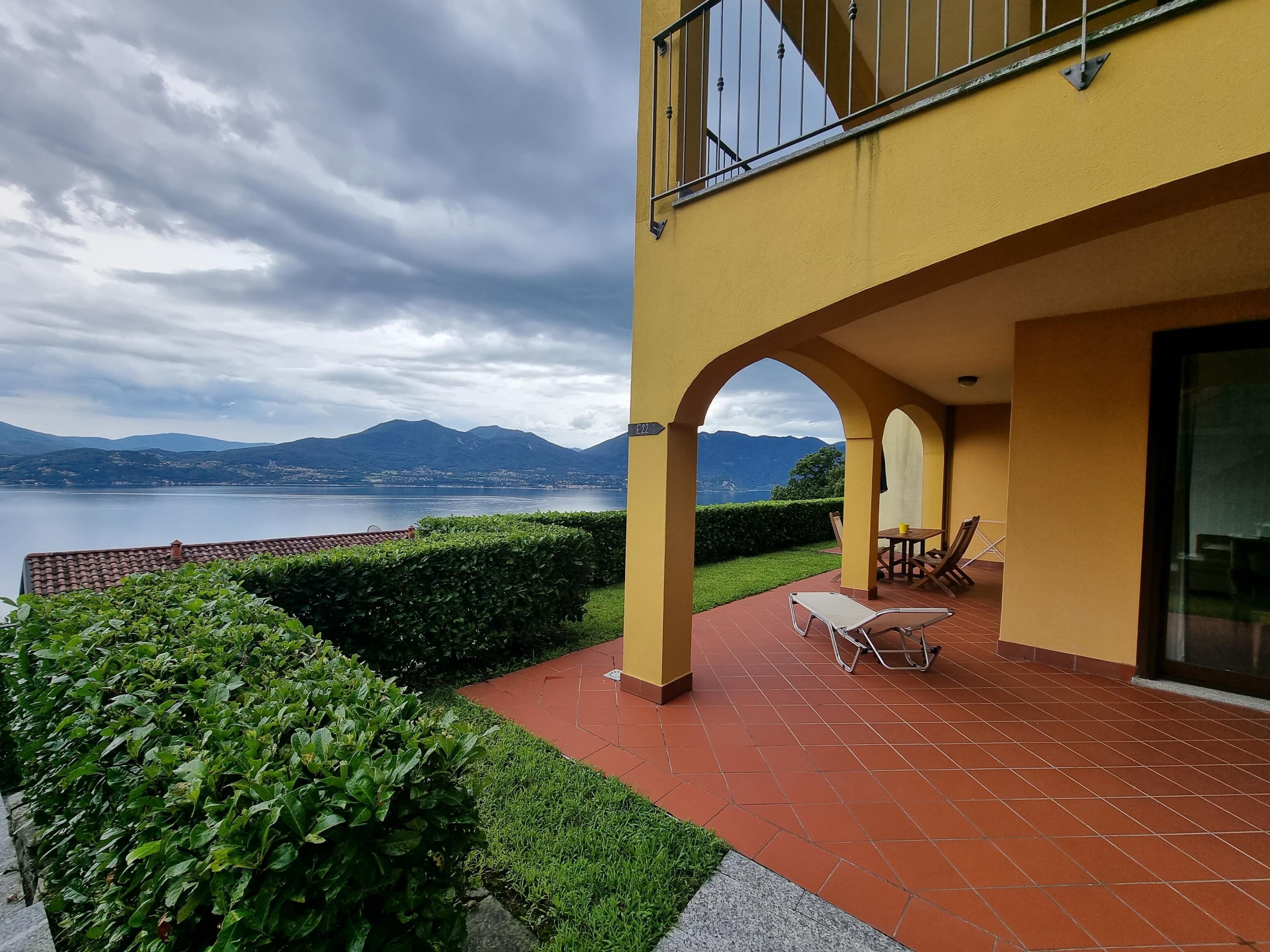 A terrace with a view of a lake and mountains, a yellow building, and a table with chairs.