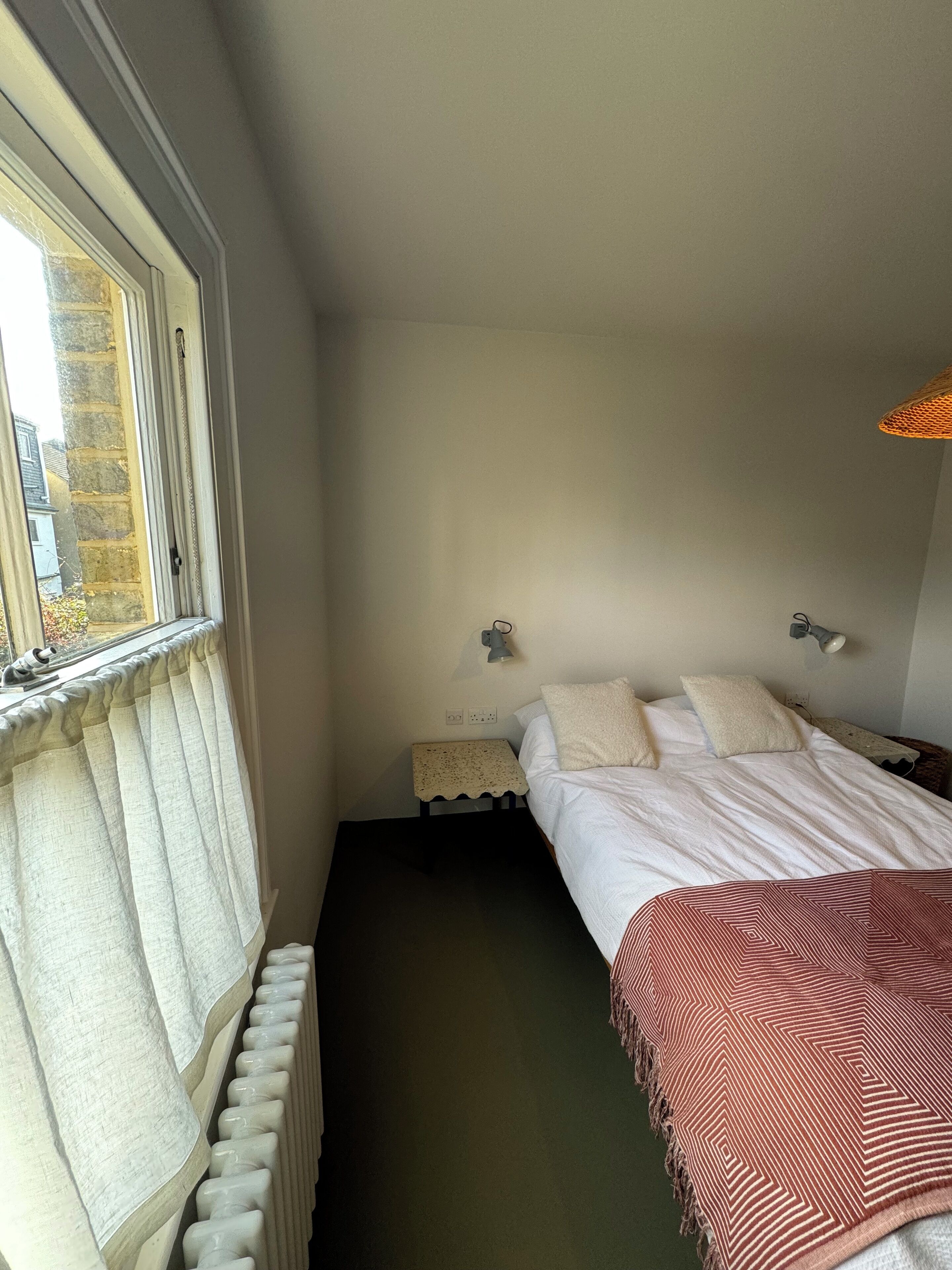 A narrow room with a bed, a small table, a lamp, and a radiator.
