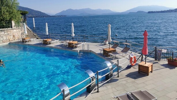 Ein Schwimmbecken mit Liegestühlen und Sonnenschirmen, umgeben von einer Steinterrasse mit Blick auf ein Gewässer und Berge im Hintergrund.