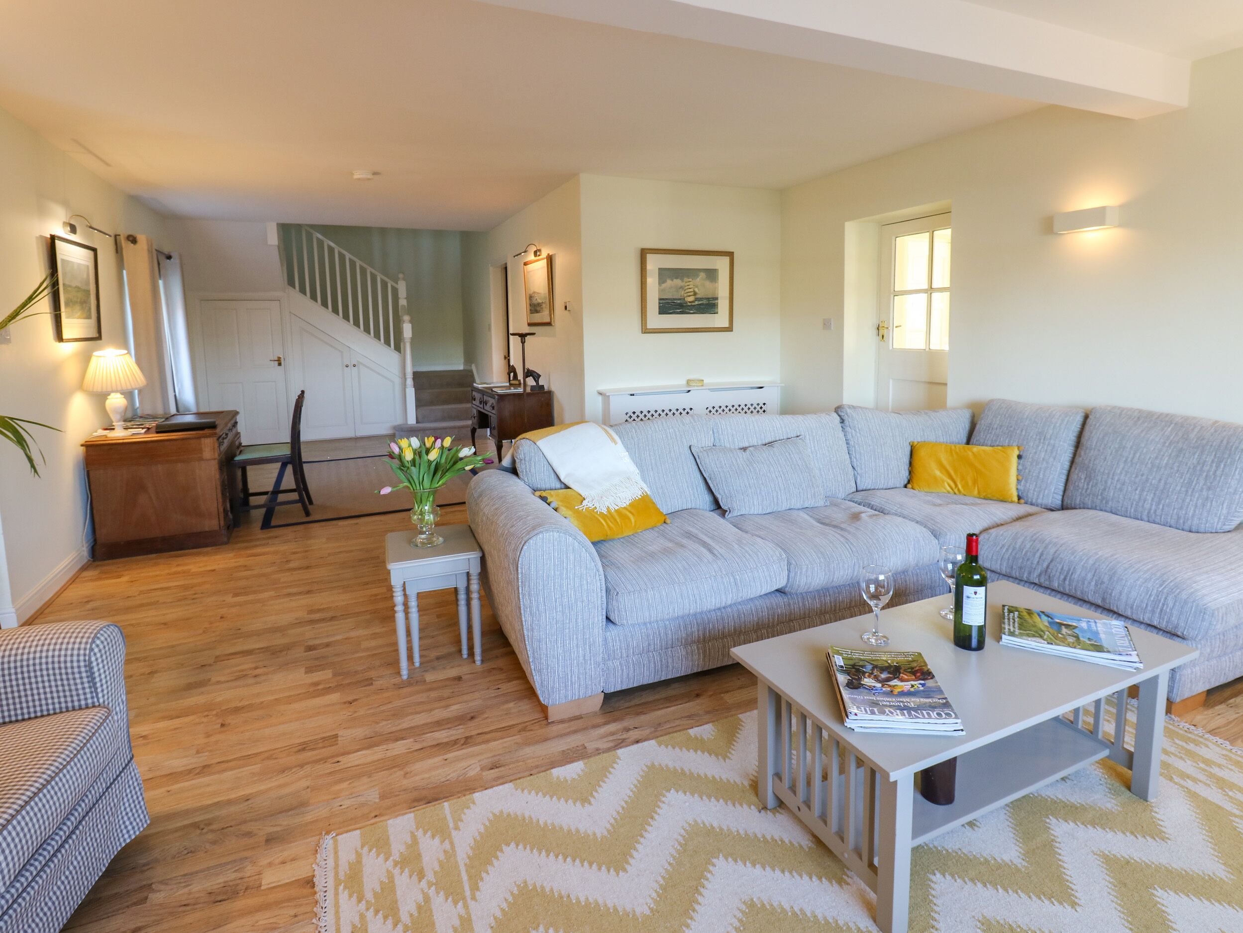 Ein Wohnzimmer mit grauem Sofa, einem kleinen Tisch mit einer Zeitschrift, einer Lampe und einer Vase mit Blumen. Im Hintergrund ist eine Treppe zu sehen.
