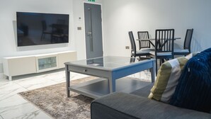 A modern hotel room with a glass-top table, a sofa, a flat-screen TV, and a dining area with chairs.