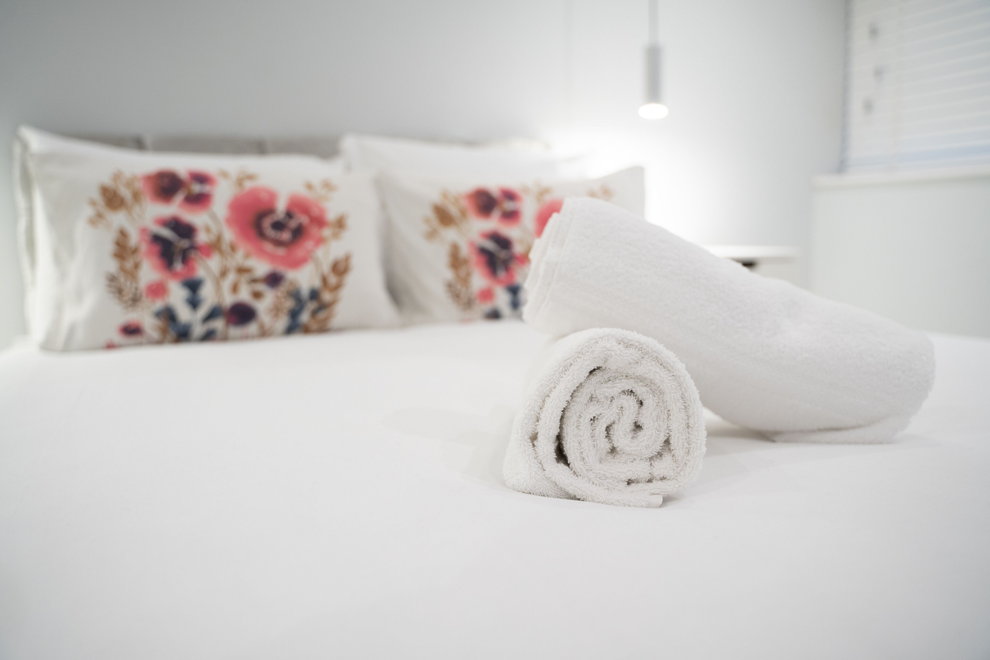 Ein ordentlich bezogenes Bett mit einem aufgerollten weißen Handtuch und blumigen Kissen.