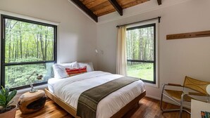 Ein Schlafzimmer mit einem großen Bett, zwei Fenstern mit Blick auf den Wald, einer Holzbalkendecke und einem kleinen Schreibtisch mit Stuhl.