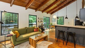 Ein geräumiger Wohnbereich mit Holzboden, große Fenster mit Blick auf den Wald, ein grünes Sofa, ein Holztisch und eine Küchenzeile mit Barhocker.