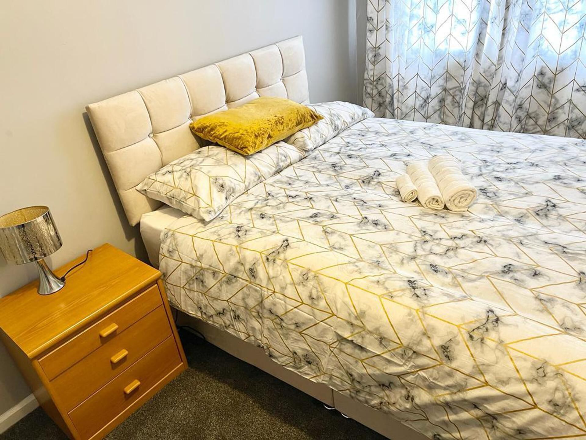 Un lit bien fait, recouvert d’une couvre-lit à motifs de marbre, avec un seul oreiller jaune et des serviettes enroulées posées sur le lit. Une table de chevet en bois, sur laquelle se trouve une lampe, est placée à côté du lit.