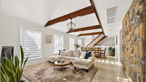 A spacious living room with a high ceiling, exposed wooden beams, a stone accent wall, a large sectional sofa, and a dining area with a staircase. - 3-bedroom vacation home just 12 minutes from Lafayette and Lehigh College. (Nazareth)