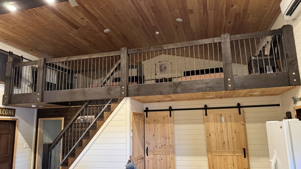 Ein geräumiger Raum mit hoher Decke, einer Holztreppe, einem Essbereich mit Tisch und Stühlen sowie einer Küchenzeile mit Kühlschrank.