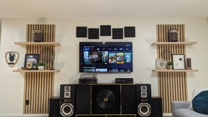 A modern living room with a flat-screen TV mounted on the wall, wooden shelves with decorative items, a large black speaker system, and a grey sofa. - Prime Downtown Music Lovers Experience! Nostalgic Mid-Mod In Historic St Peter (Saint Peter)