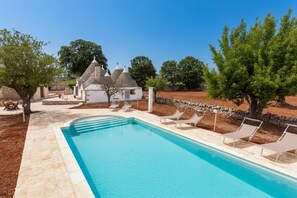 Una piscina con sillones de descanso, un edificio de piedra y árboles al fondo.