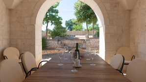 Une table en bois, dressée pour un repas, avec des chaises, une bouteille de vin et des verres, dans un cadre extérieur agrémenté de murs en pierre et d’arbres.