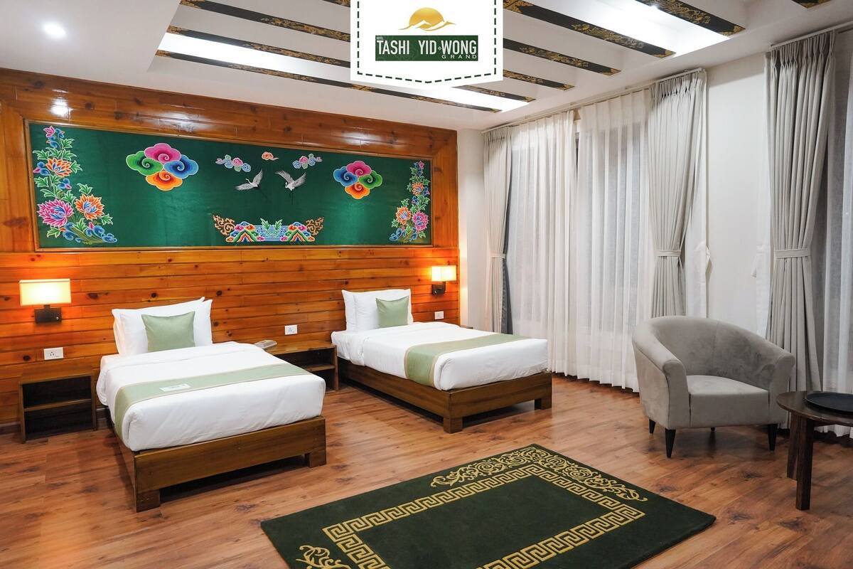 A hotel room with two beds, wooden headboard, a green wall with floral and cloud artwork, a grey armchair, and a green patterned rug.