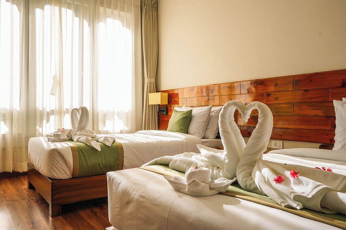 A hotel room with two beds, wooden headboard, white bedding, and a heart-shaped towel decoration.