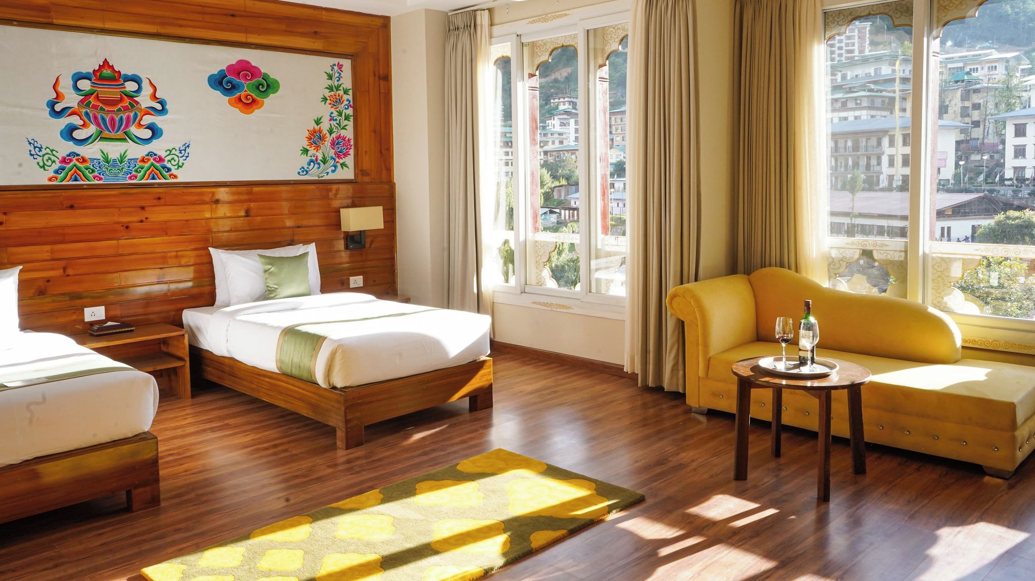 A hotel room with a wooden headboard, two beds, a sofa, a small table, and a view of the city through large windows.