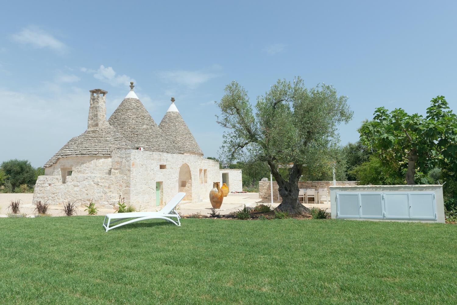 A stone building with conical roofs, a tree, and a white lounge chair in a grassy area.