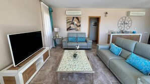 Un salón con una televisión de pantalla plana, dos sofás grises con cojines azules, una mesa de centro con motivos y un reloj de pared decorativo.