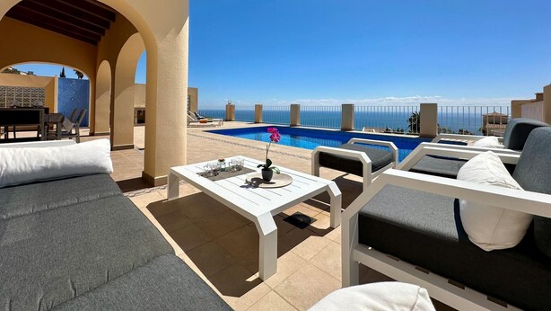 Ein Poolbereich mit Liegestühlen, einem Tisch und Blick auf das Meer.