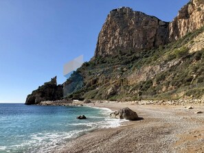 Une plage rocheuse bordée d’une eau d’un bleu limpide, une grande formation rocheuse et une falaise abrupte.