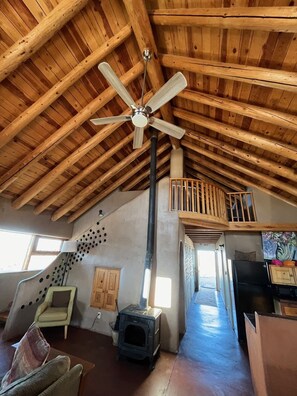 A spacious loft with a high wooden ceiling, a central ceiling fan, a wood-burning stove, a dining area with a table and chairs, and a kitchen with a refrigerator. - Special Rates-Private Taos Home. Beautiful Oasis. (El Prado)