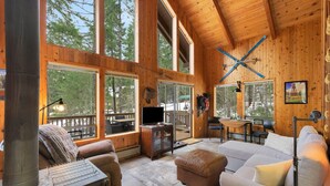 Un interior de cabaña acogedor con chimenea, ventanas amplias, televisor y zona de comedor.