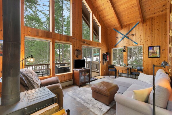 Un interior de cabaña acogedor con chimenea, ventanas amplias, televisor y zona de comedor.