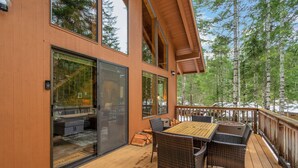 Una terraza de madera con una mesa y sillas, un gran ventanal corredero y vistas a un bosque.