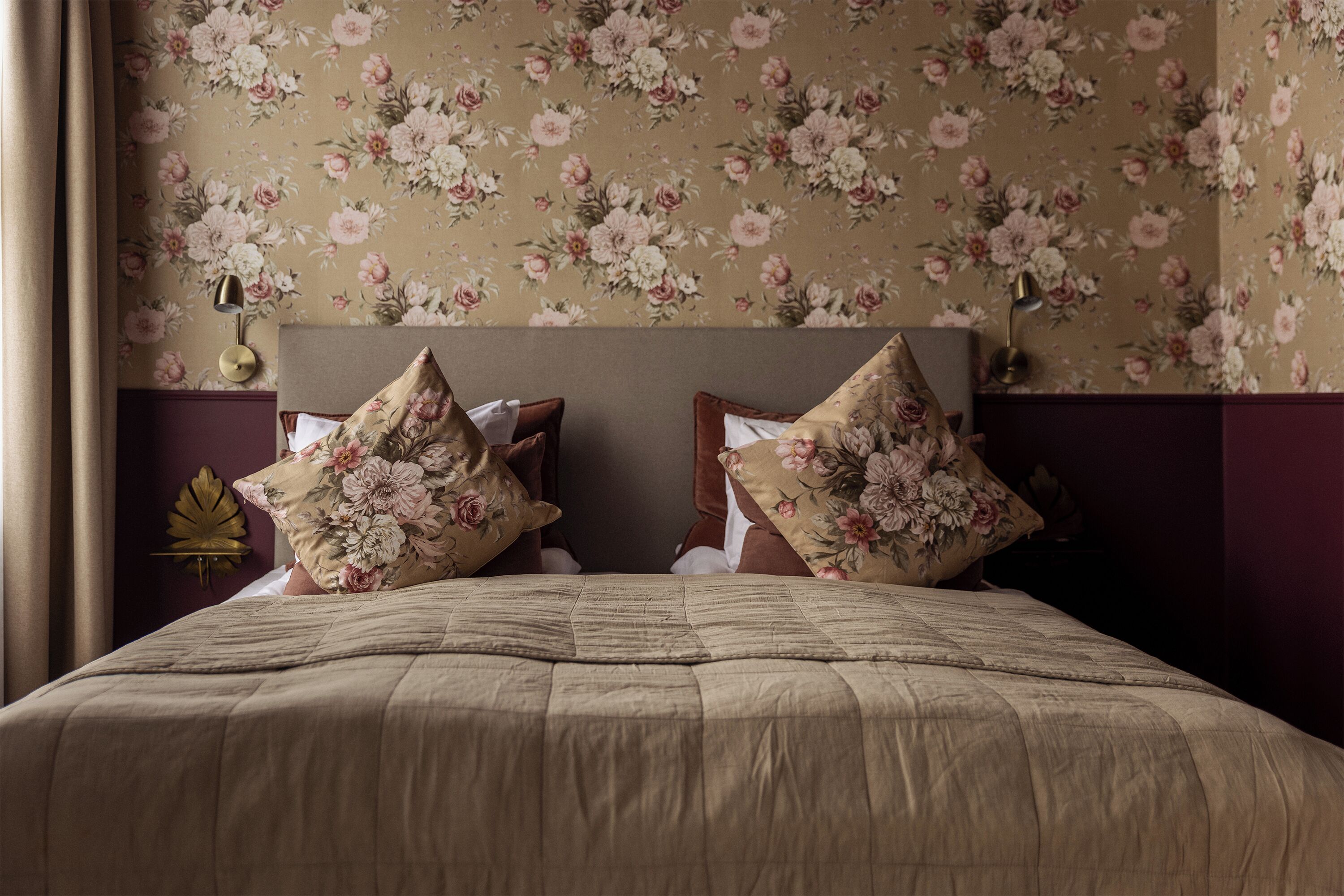 Una cama con almohadas estampadas con motivos florales y una colcha color beige en una habitación con papel tapiz floral y un cabecero de madera oscura.