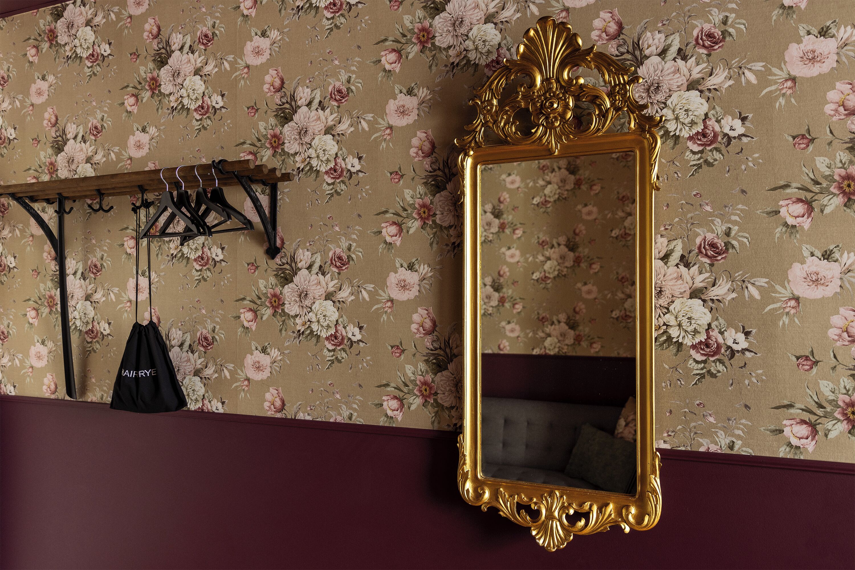 Una habitación con papel tapiz floral, un espejo con marco dorado y un bolso negro colgado en un perchero.