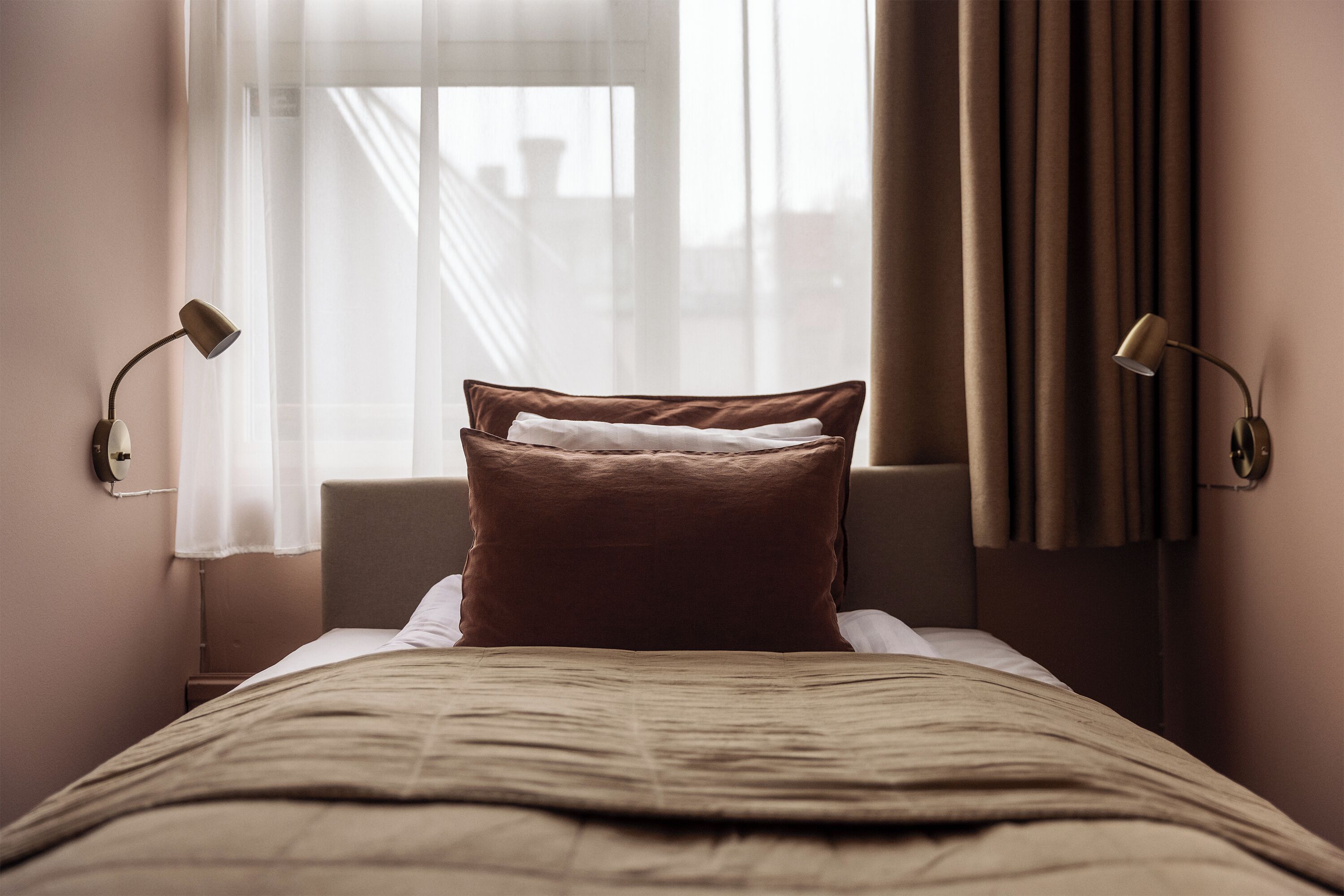 Una cama bien hecha con un cabecero marrón, dos lámparas de noche y una ventana con cortinas traslúcidas.