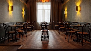 A dining area with wooden flooring, round tables, and chairs, featuring a central chandelier and wall sconces.