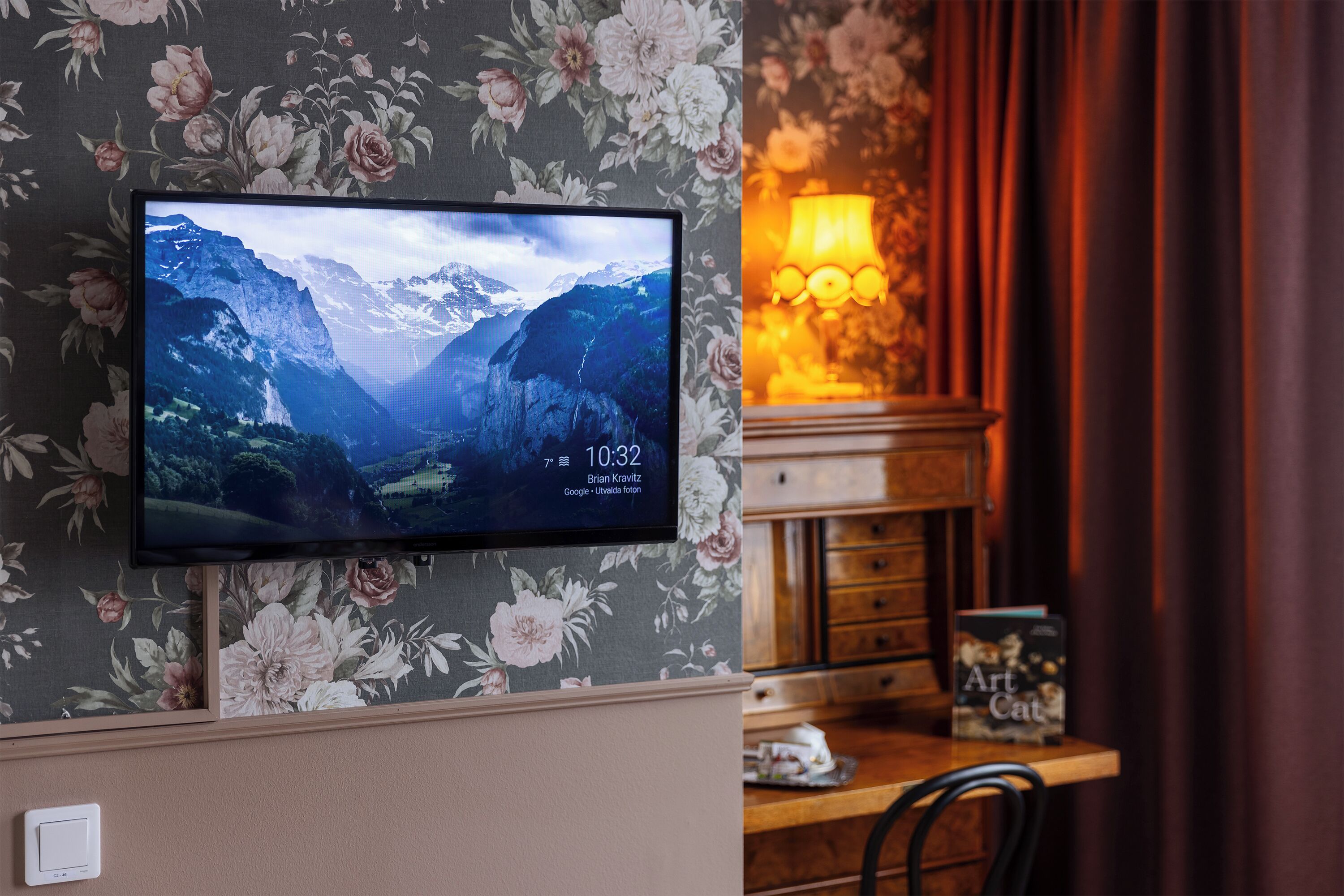 A television mounted on a floral wallpaper wall displaying a mountainous landscape.