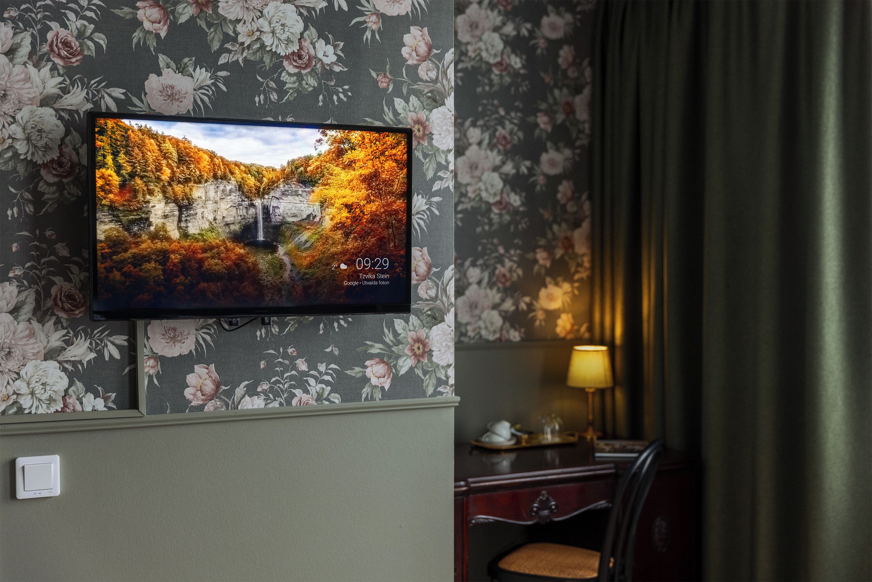 A room with a TV displaying a scenic autumn landscape, a desk with a lamp, and floral wallpaper.