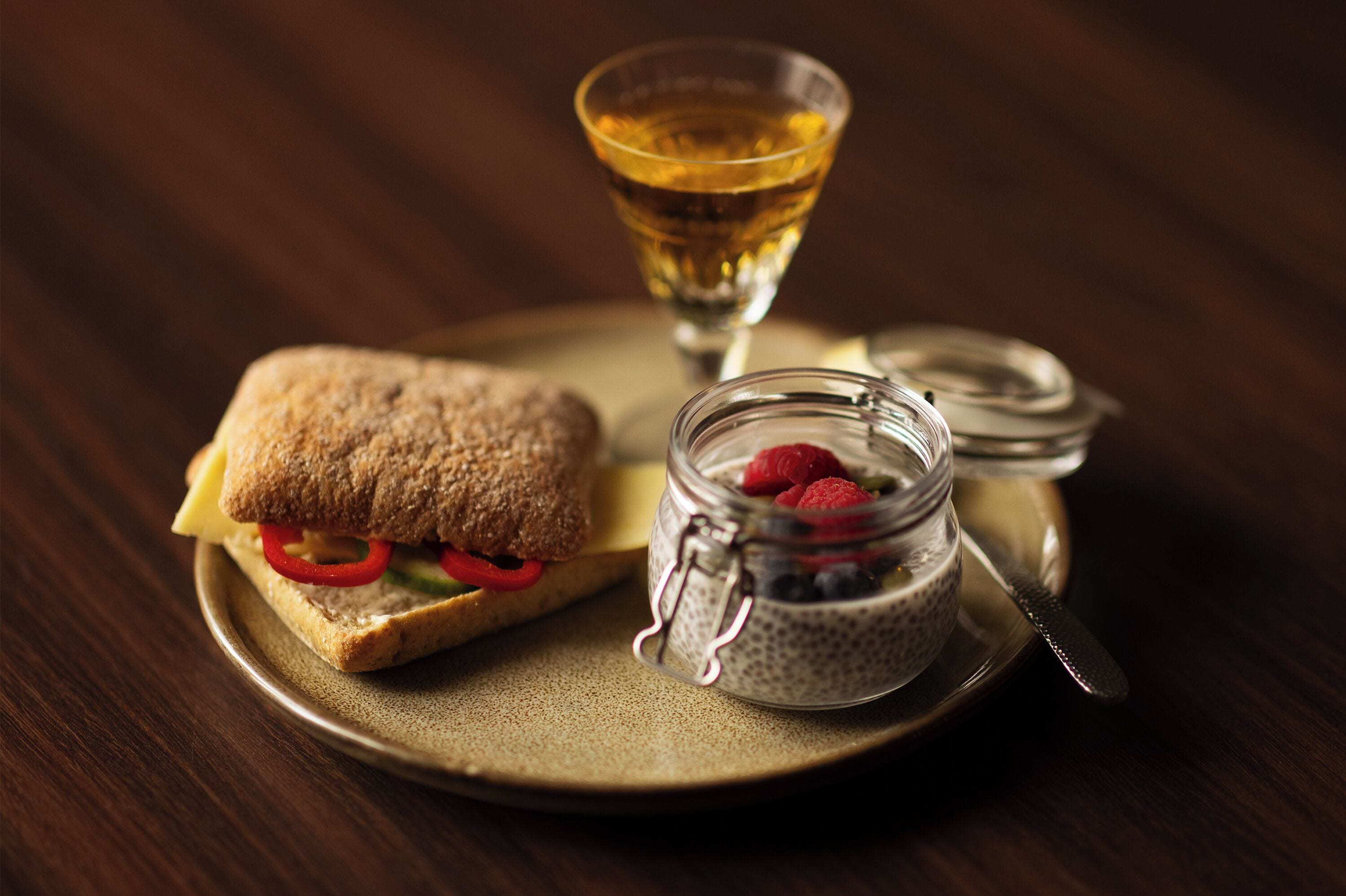 A sandwich with a glass jar of chia seeds and raspberries, and a glass of amber liquid on a plate.
